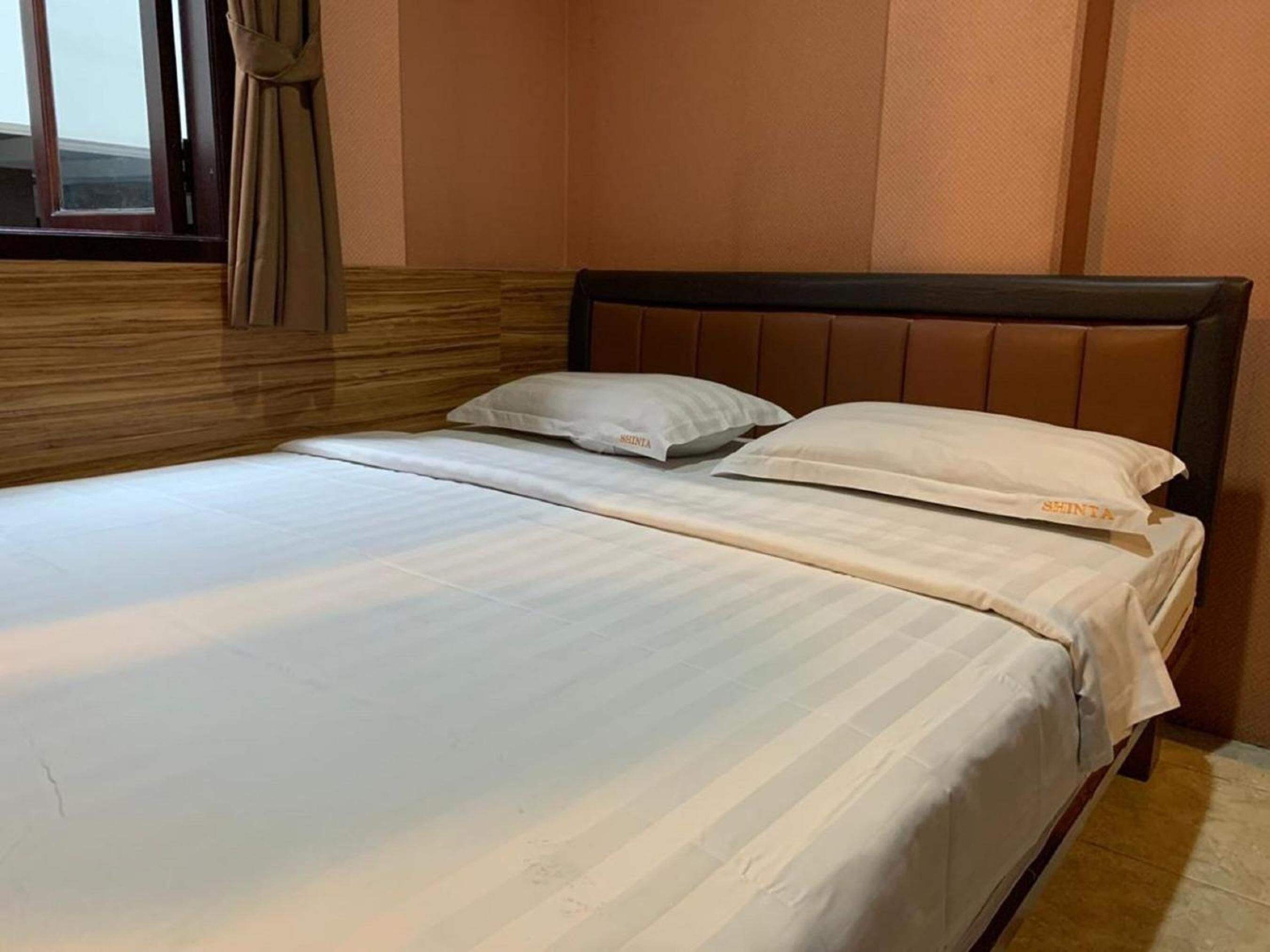 A neatly made bed with white linens and pillows, a wooden headboard, and a window with curtains.