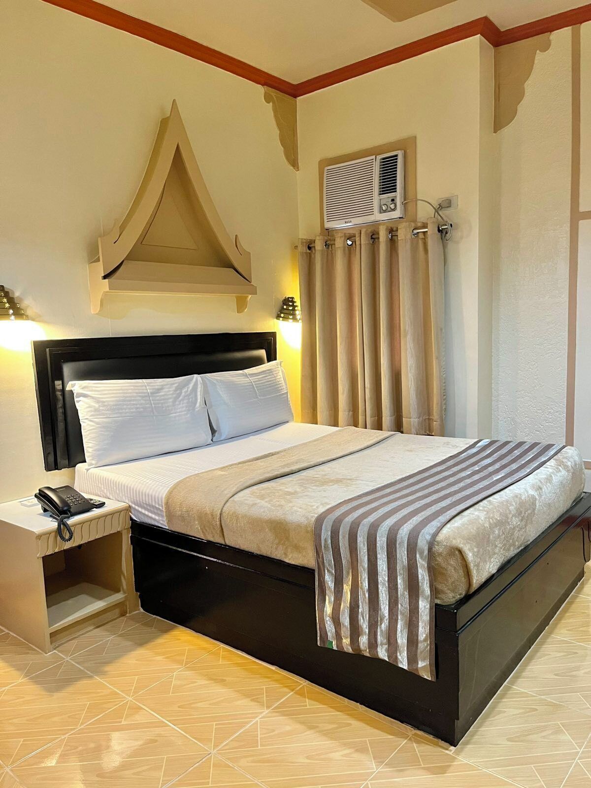 A hotel room with a bed, bedside table, and an air conditioning unit.