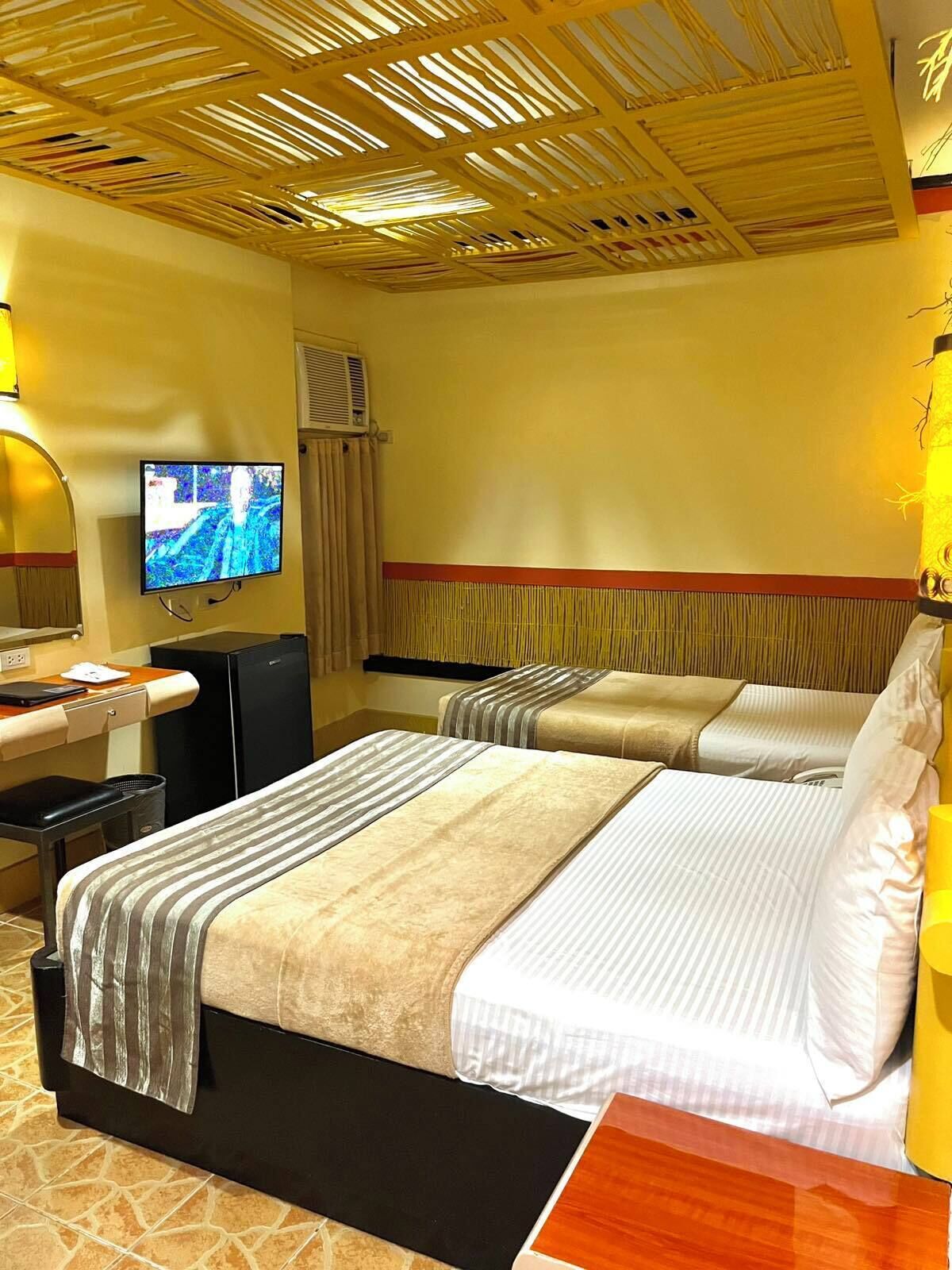 A hotel room with two beds, a desk with a computer, a television, and a ceiling with a unique design.