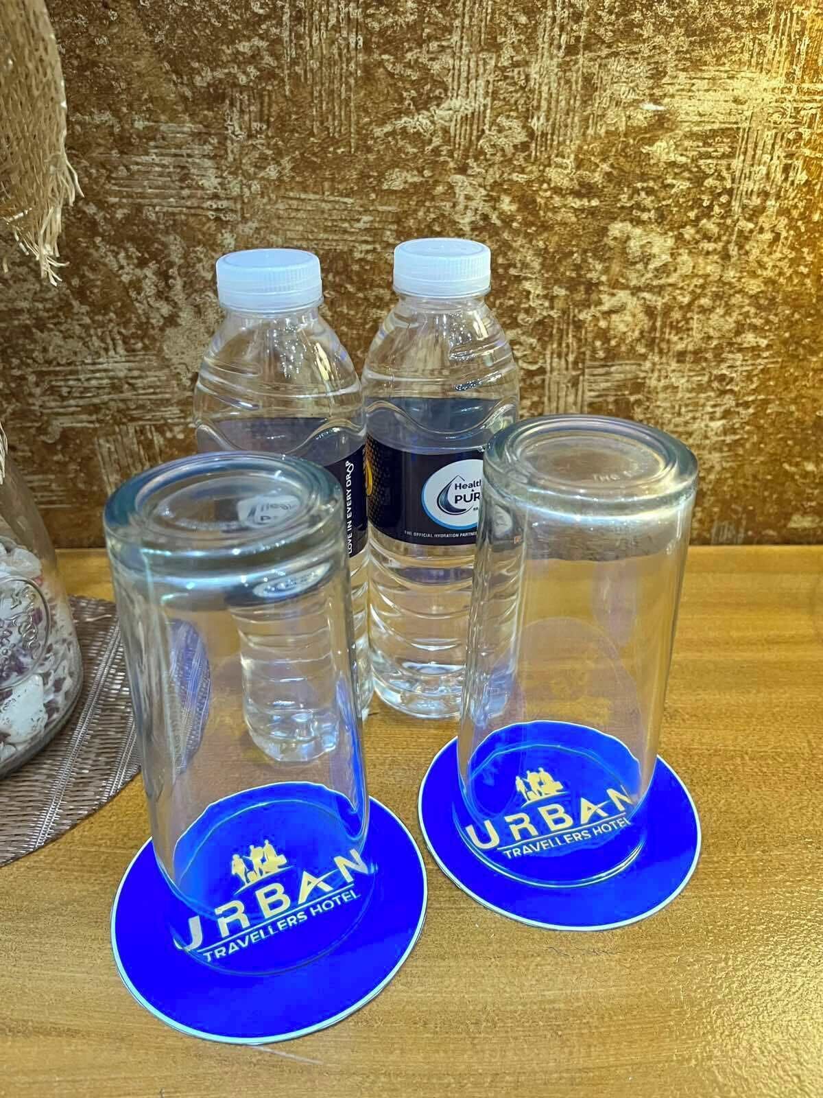 Trois bouteilles en verre transparent et deux verres en verre transparent, accompagnés de deux sous-verres bleus, dont l’un porte l’inscription « URBAN TRAVELERS HOTEL ».