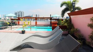 Un área de piscina en la azotea con sillones de descanso, una piscina y un horizonte urbano de fondo.