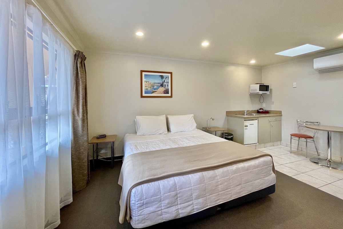 Quarto de hotel com cama, escrivaninha, cadeira, mesinha, pia e micro-ondas.
