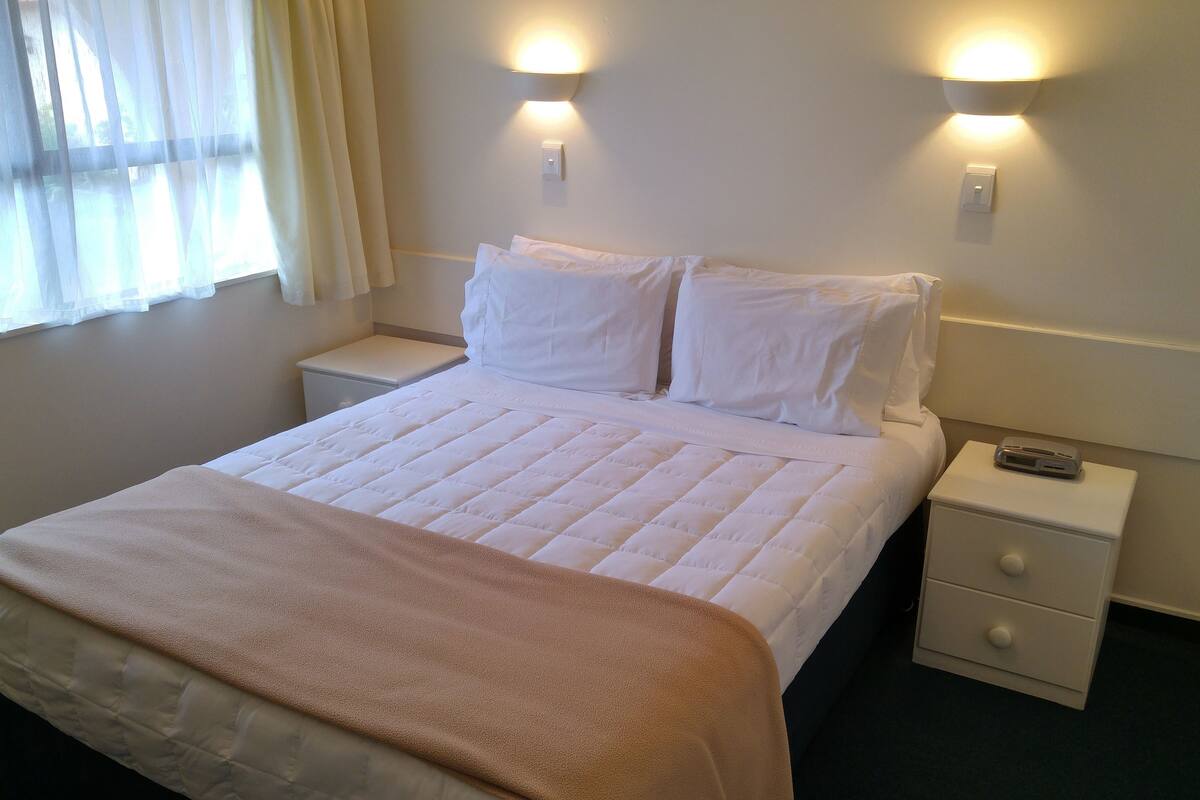 Uma cama bem arrumada com edredom branco e manta marrom, duas mesas de cabeceira com abajures, uma janela com cortinas leves e um luminário fixado na parede.