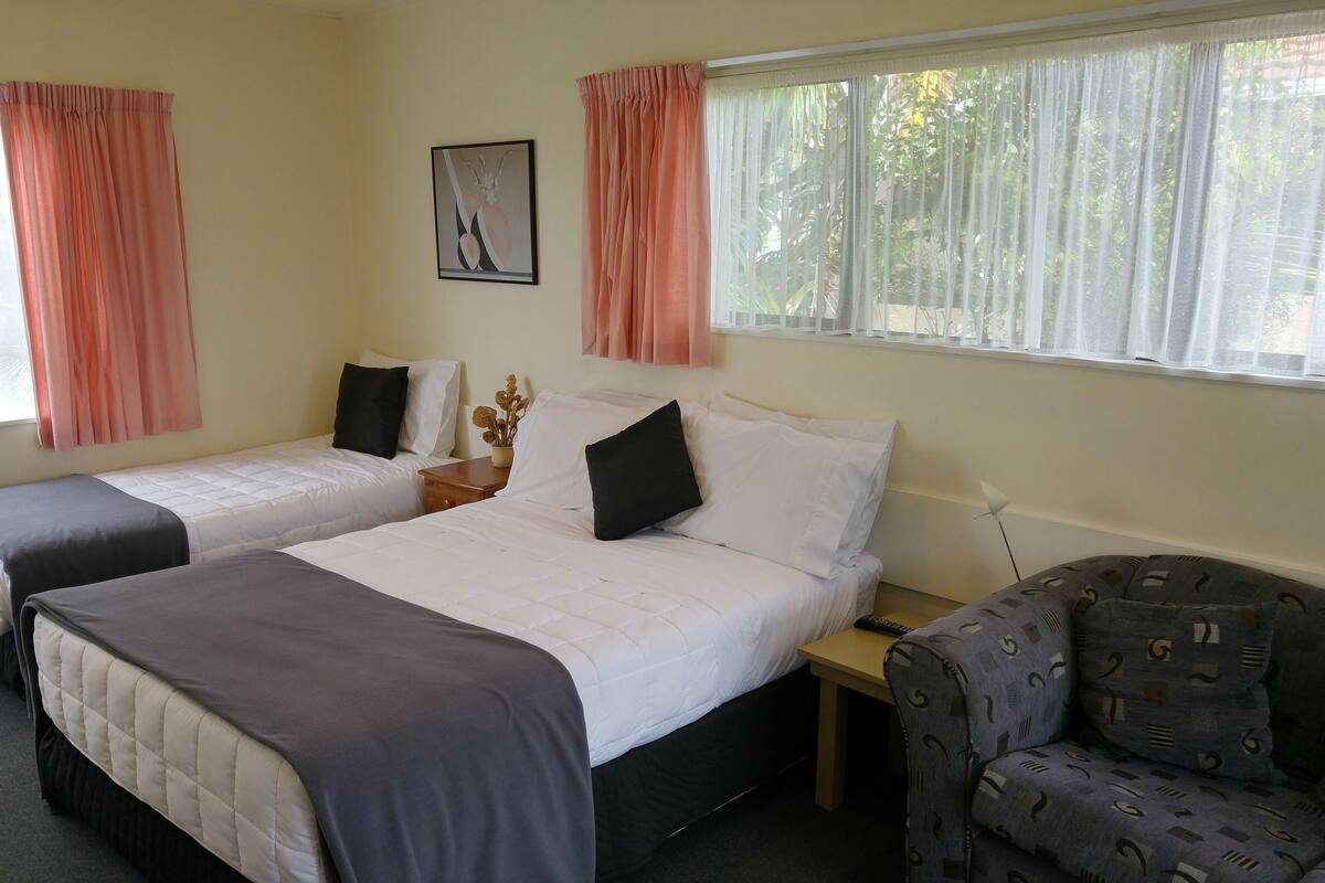 Quarto de hotel com duas camas de solteiro, uma mesinha, uma cadeira e uma janela com cortinas.