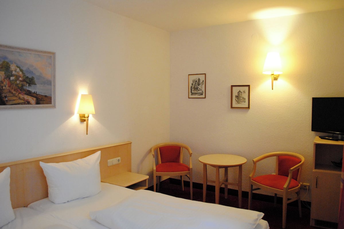 Habitación de hotel con cama, dos sillas, una mesita, televisor y lámparas de pared.