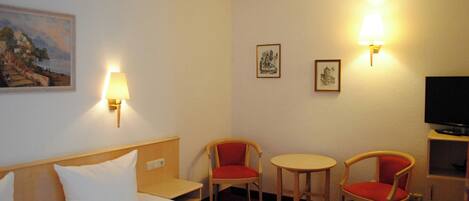 Habitación de hotel con cama, dos sillas, una mesita, televisor y lámparas de pared.