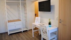 Habitació amb escriptori blanc, cadira, armari blanc amb una banca i televisió penjada.
