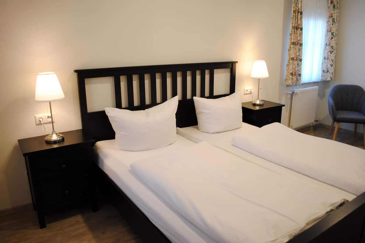 Una cama doble con ropa blanca, dos mesitas de noche con lámparas, una silla y una ventana con cortinas estampadas.