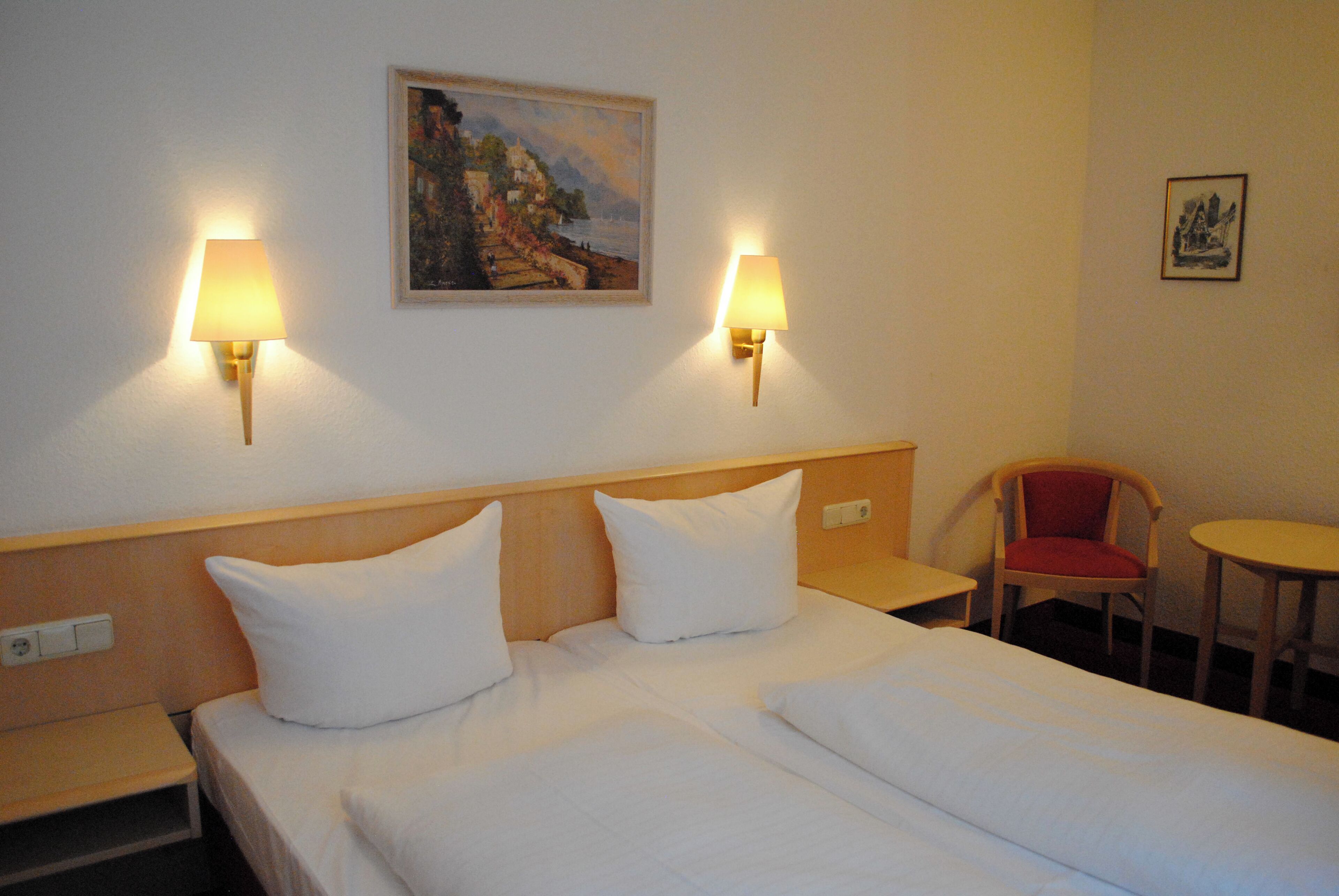 A hotel room with two beds, two bedside lamps, a painting on the wall, a chair, and a small table.