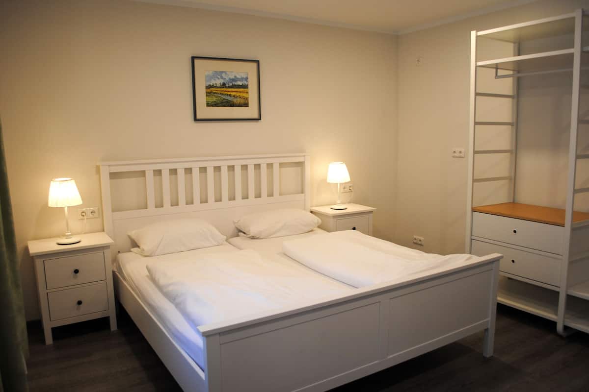 Una cama bien hecha con ropa blanca, dos mesitas de noche con lámparas, un cabecero blanco, un estante montado en la pared y un cuadro enmarcado sobre la cama.