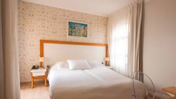 A hotel room with a bed, a nightstand with a phone, a lamp, a painting on the wall, and a chair.