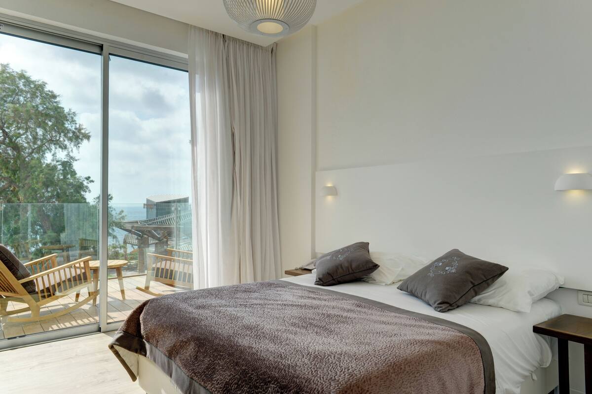 A modern hotel room with a large bed, two pillows, a bedside table, and a view of a balcony with outdoor furniture.