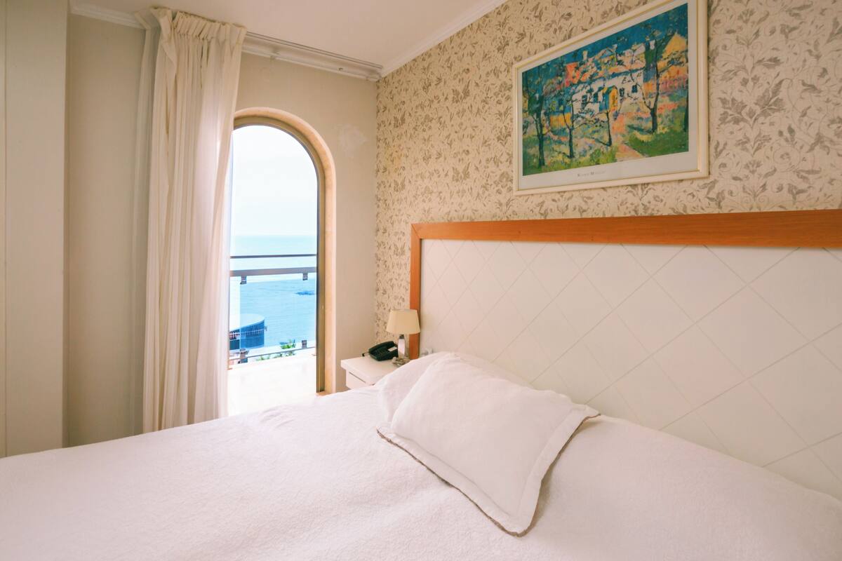 A hotel room with a bed, a nightstand, a painting, and a view of the sea through an arched window.