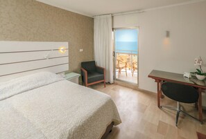 A hotel room with a bed, a desk, a chair, a small table, a curtain, and a view of the sea through a glass door. - Residence Beach (Netanya)