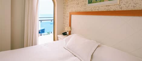 Ein Hotelzimmer mit Bett, Nachttisch, einem Gemälde und Meerblick durch ein Rundbogenfenster.
