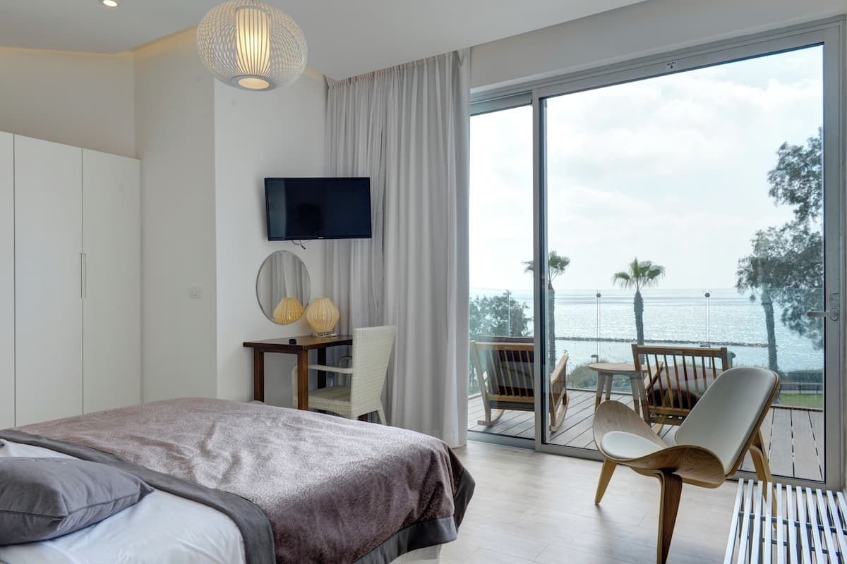 A bedroom with a bed, a TV, a desk with a chair, and a balcony with a view of the sea.