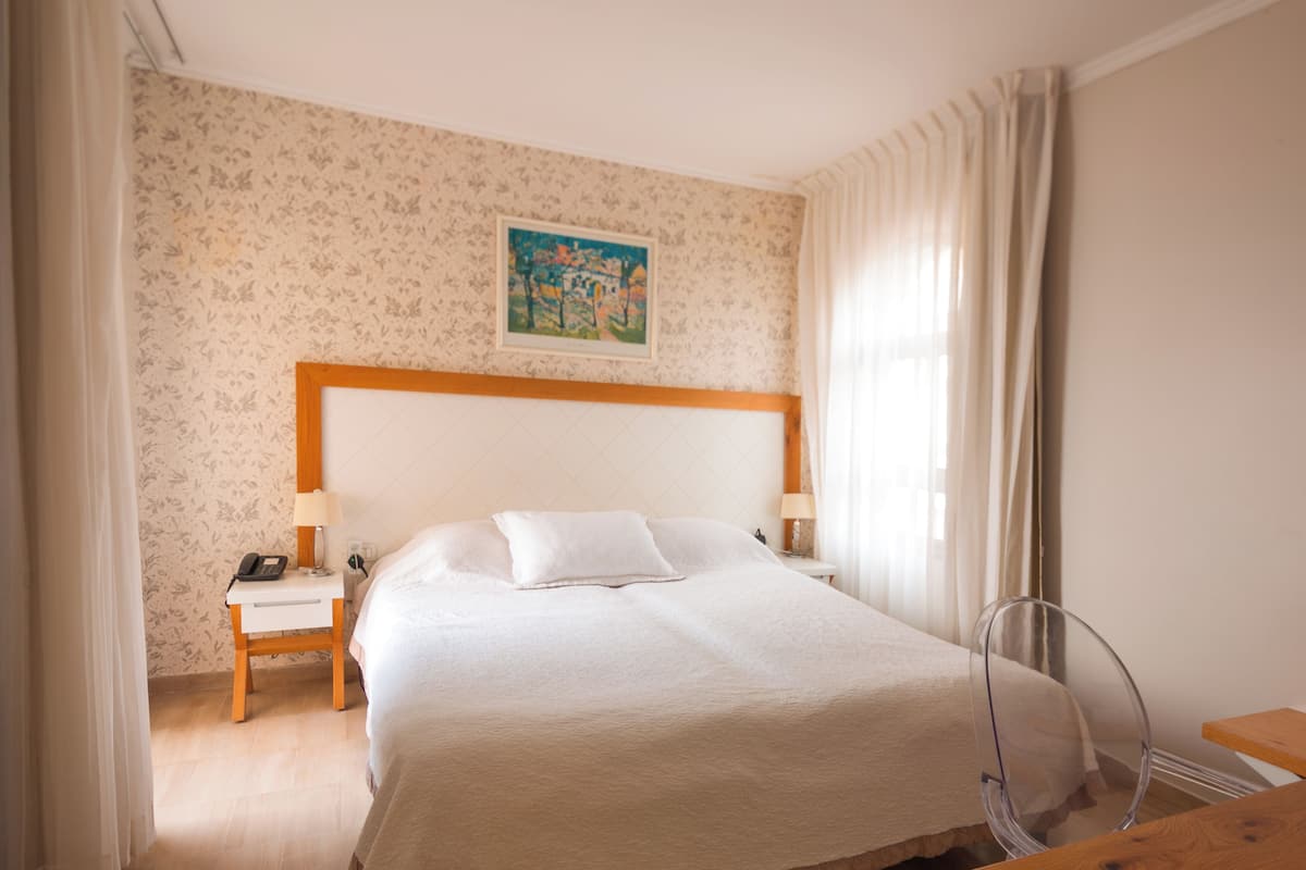 A hotel room with a bed, a nightstand with a phone, a lamp, a painting on the wall, and a chair.