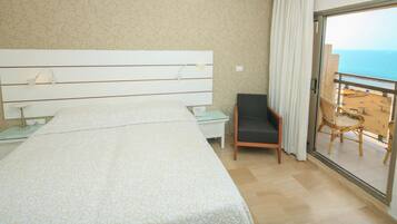 A hotel room with a large bed, a chair, a nightstand, and a balcony with a view of the beach.