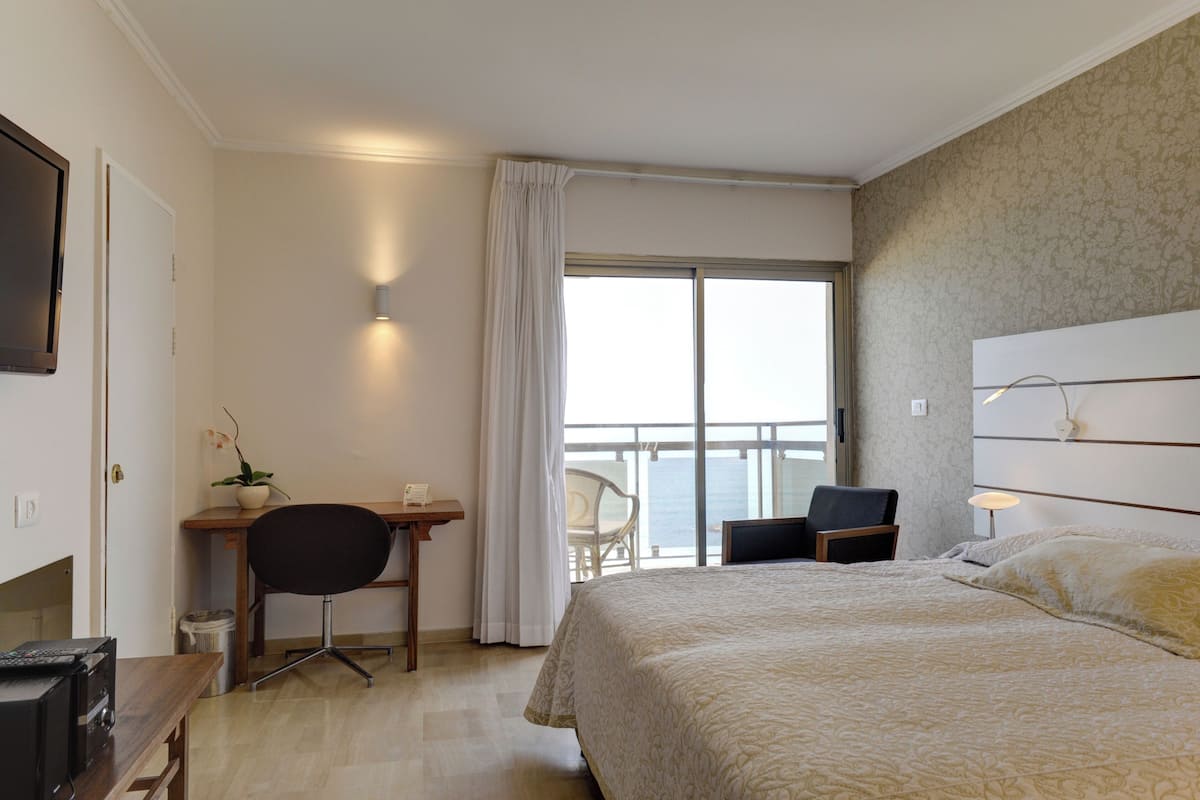 A hotel room with a bed, a desk, a chair, a TV, and a view of the sea.