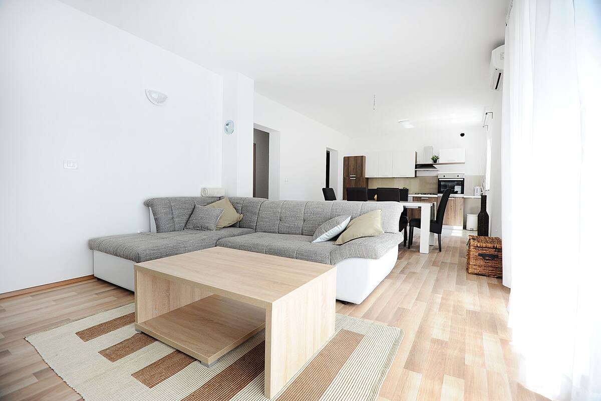 Un salon moderne avec un canapé d’angle, une table basse en bois et une cuisine en arrière-plan.