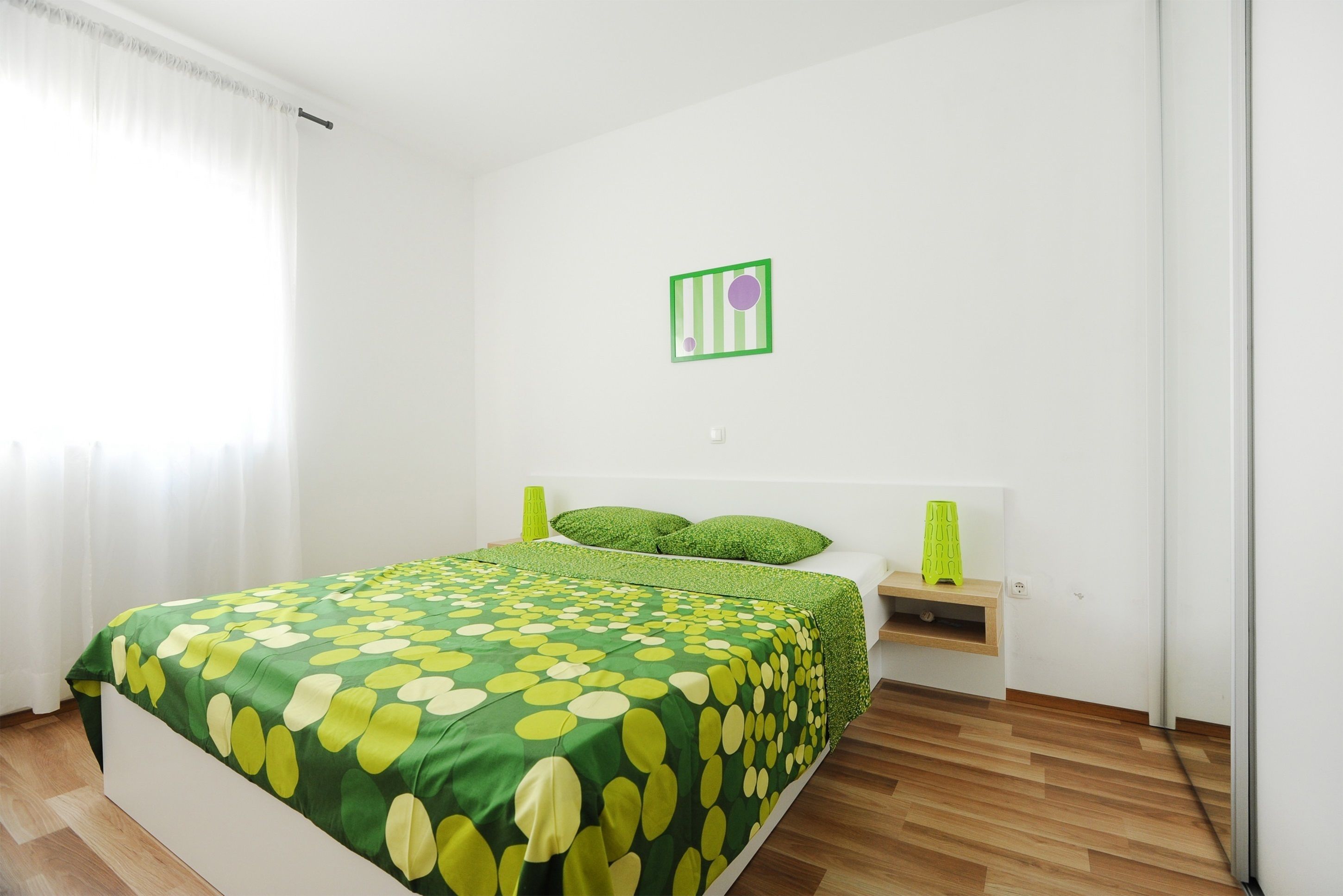 A bedroom with a bed, green pillows, a bedside table, and a green and white striped artwork on the wall.