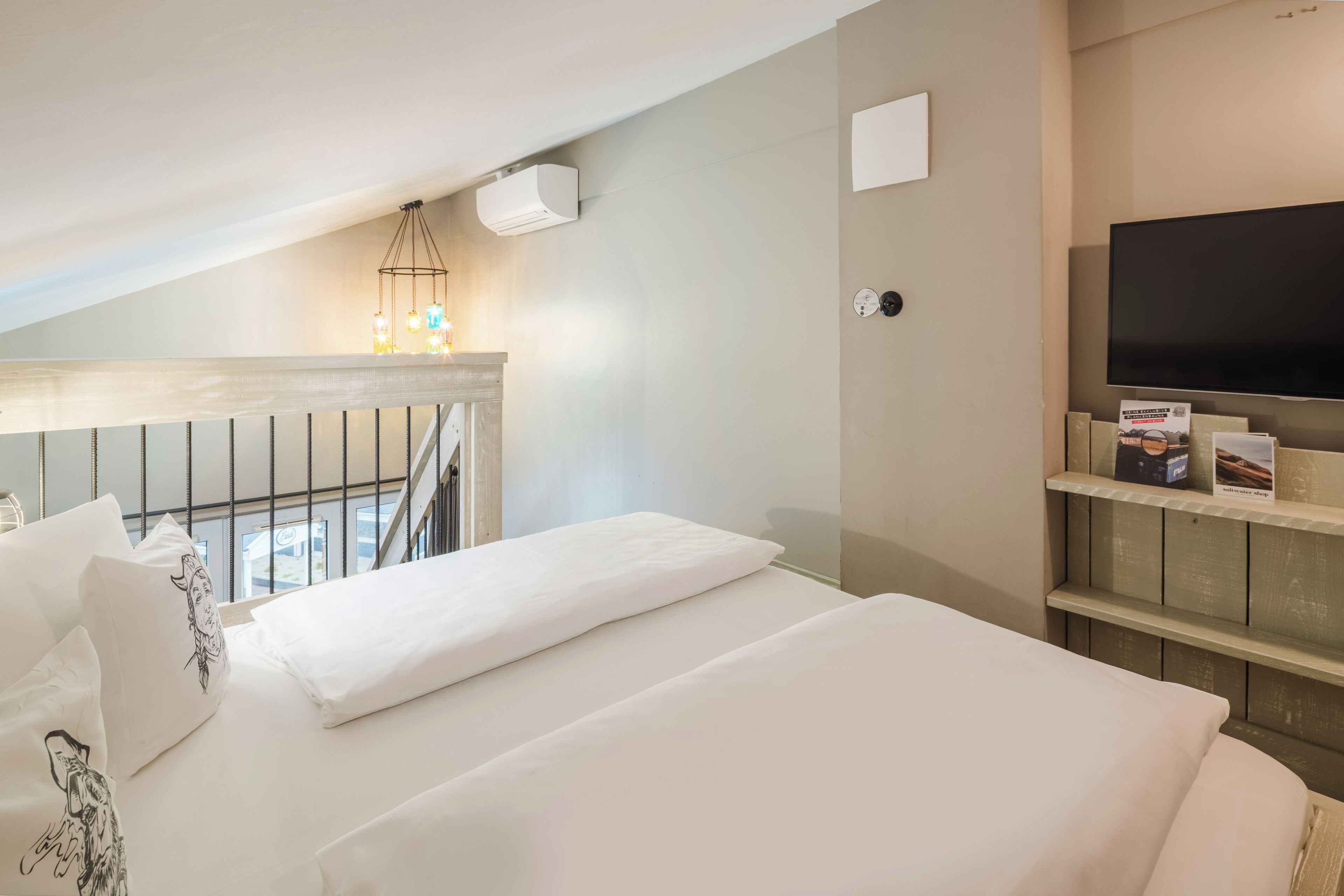 Habitación de hotel con una cama, televisor, estante con libros y balcón con barandilla.