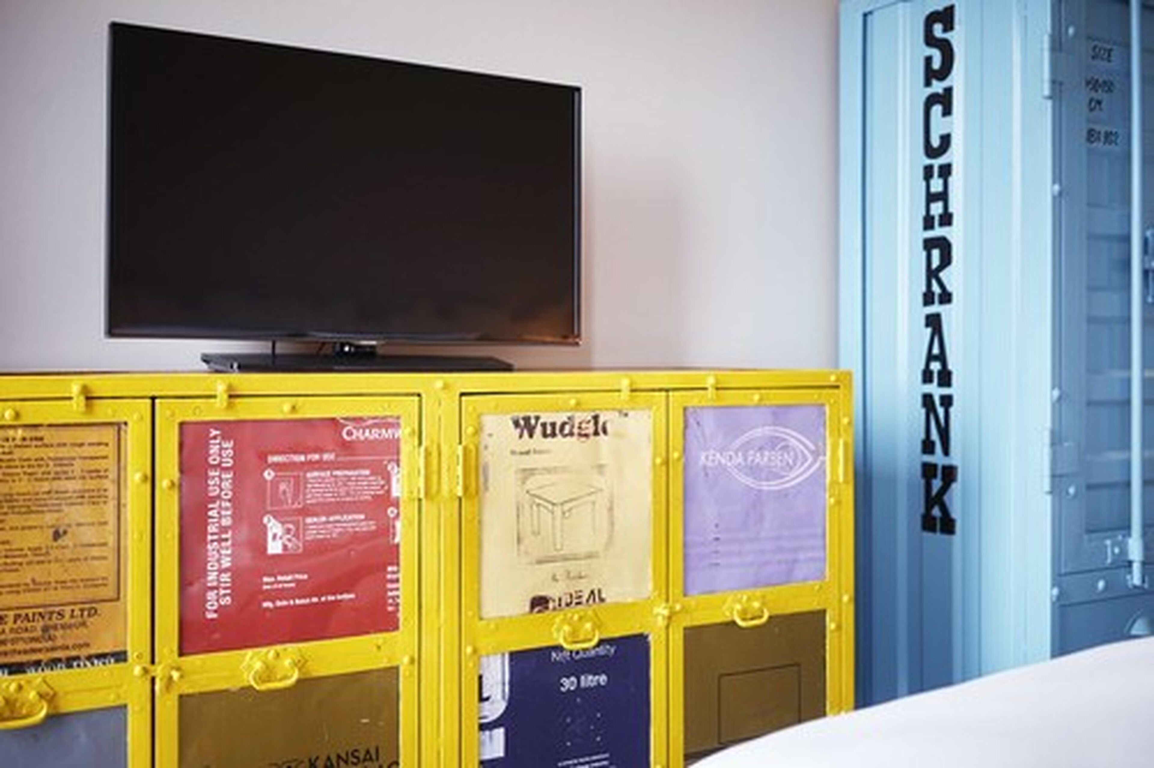A room with a television mounted on the wall, a yellow cabinet with various signs, and a blue cabinet with the word "SCHRANK" on it.