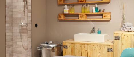A bathroom with a shower, wooden shelves with toiletries, a sink, and a towel on a bed.