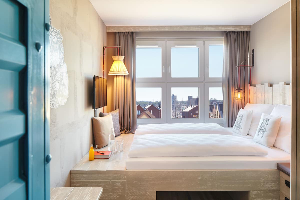 A hotel room with a large window, a bed, a bedside table with a lamp, and a view of the cityscape.