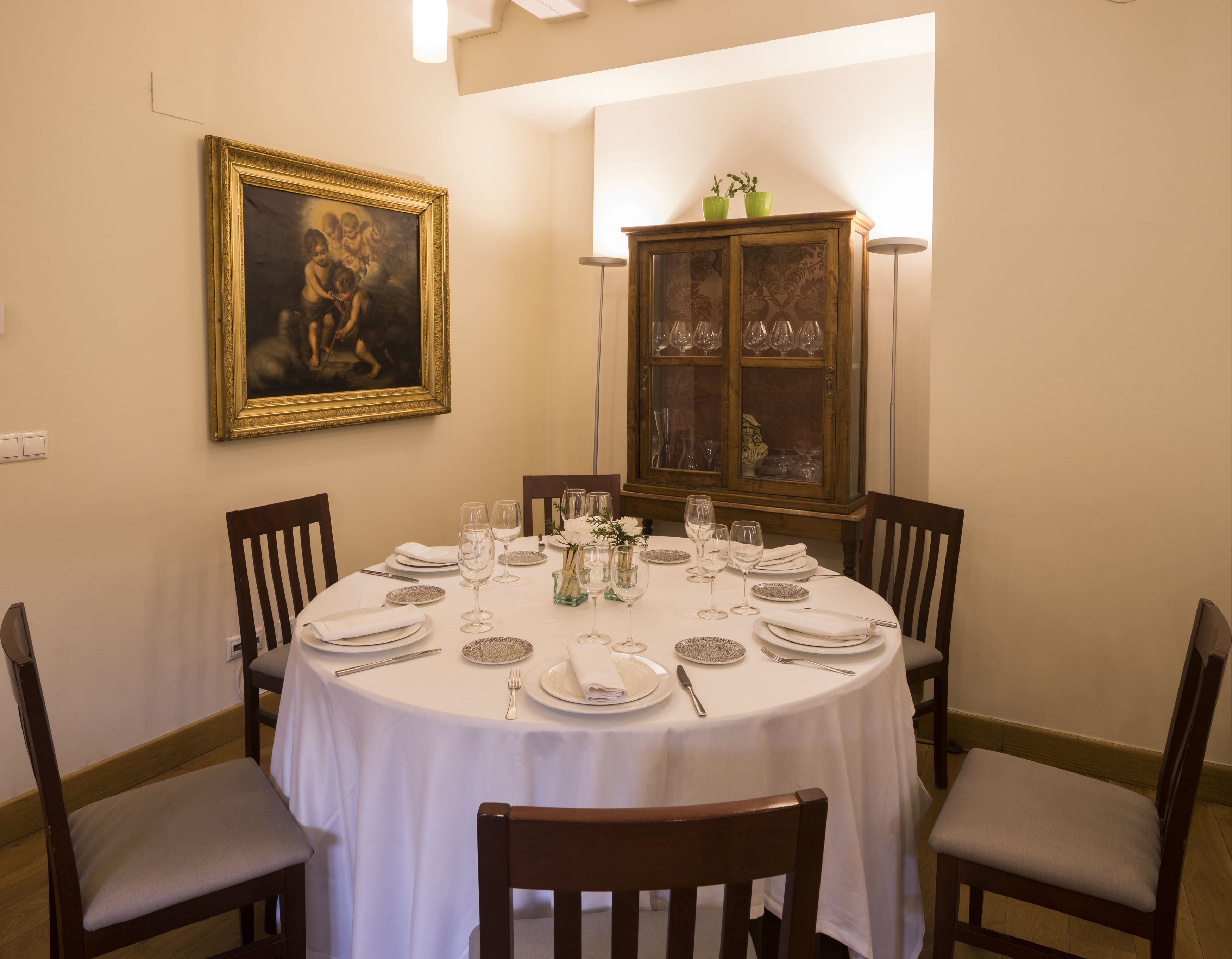 A dining room with a round table set for four, surrounded by wooden chairs. A cabinet with glass doors is visible in the background.