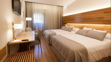 Habitación de hotel con cabecera de madera, dos camas, un escritorio con lámpara, una silla y un televisor en la pared.