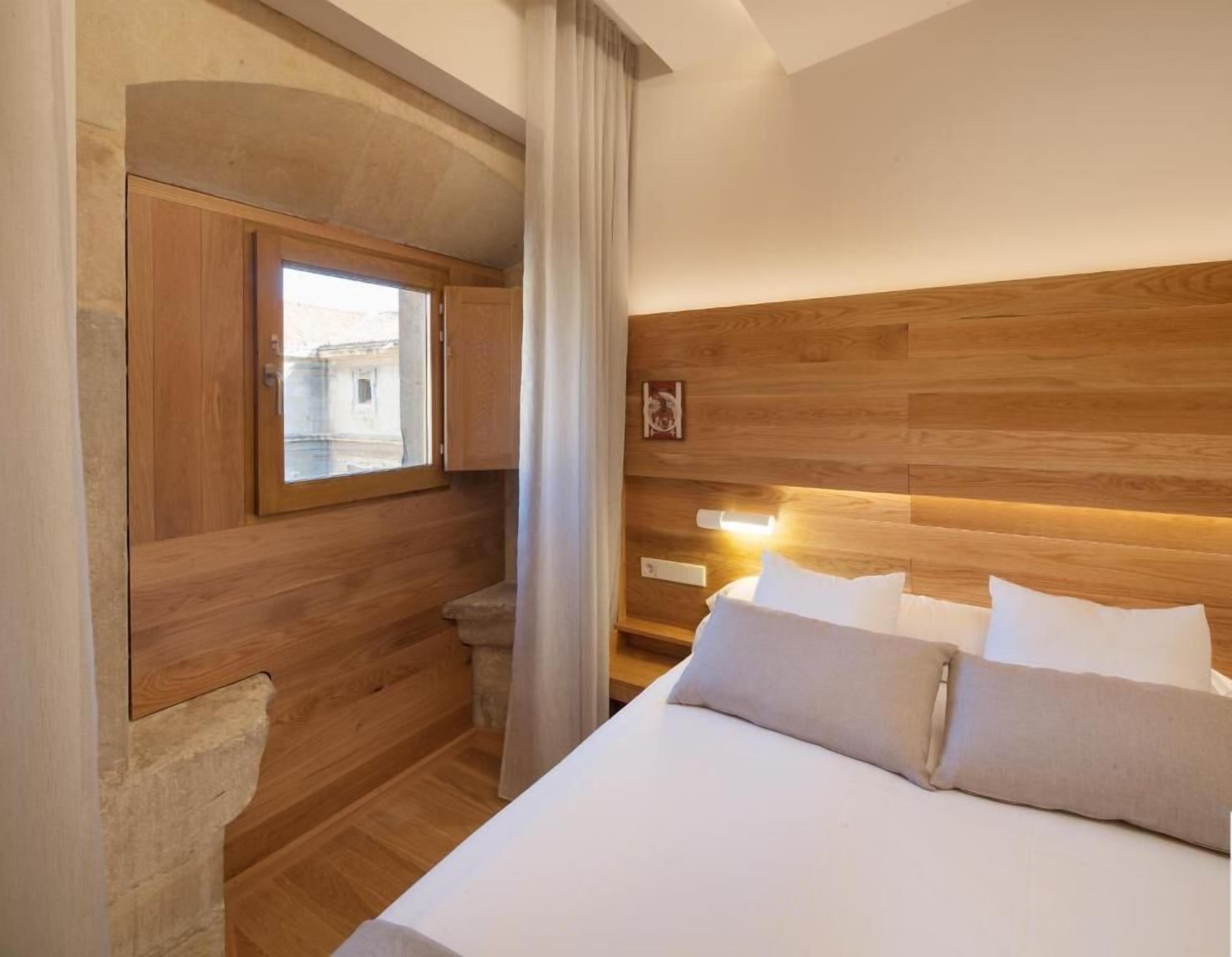 A hotel room with a wooden headboard, a bed with white linens, a window with a view of buildings, and a curtain.