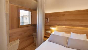 A hotel room with a wooden headboard, a bed with white linens, a window with a view of buildings, and a curtain.