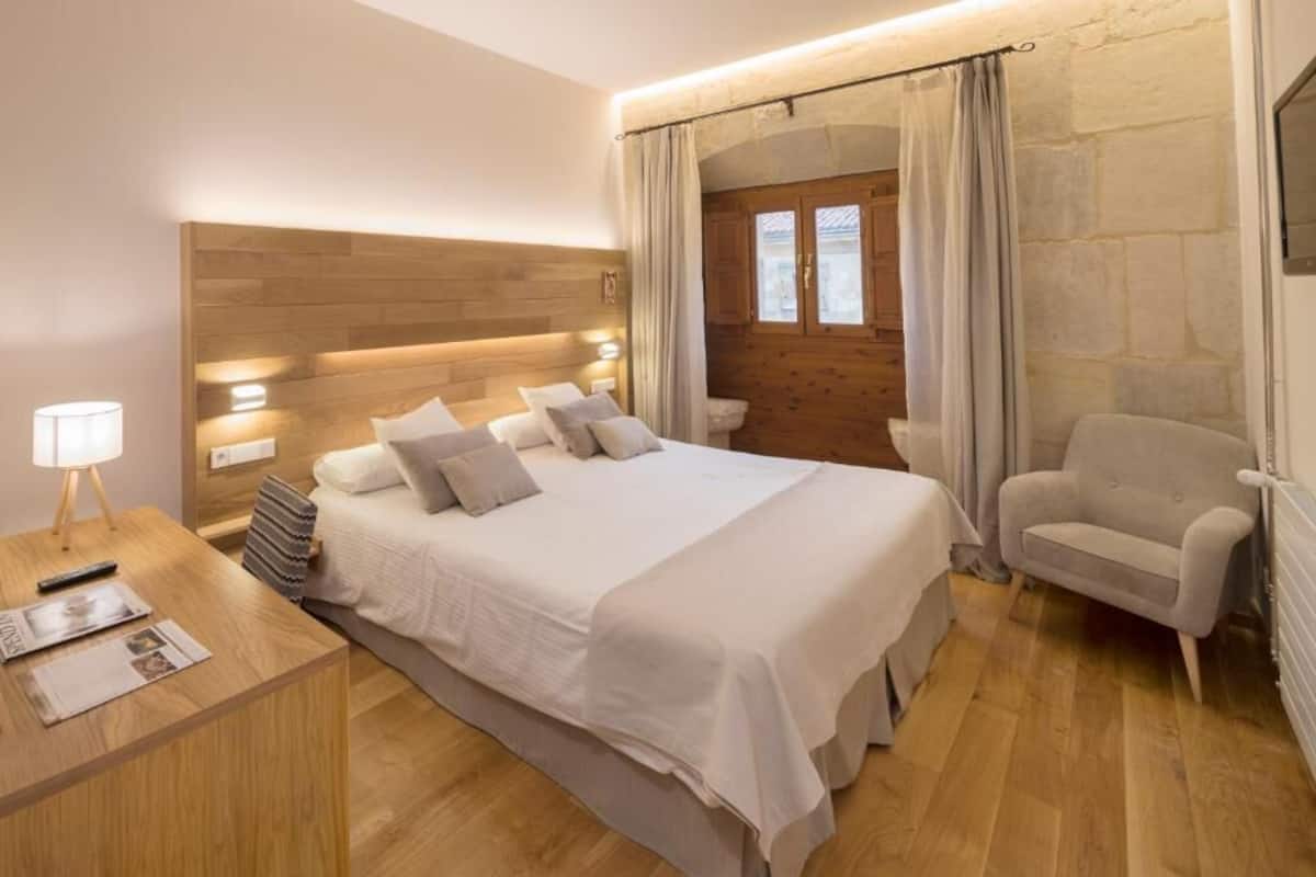 A bedroom with a wooden headboard, a bed with white bedding, a bedside table with a lamp, a chair, and a stone wall with a window.
