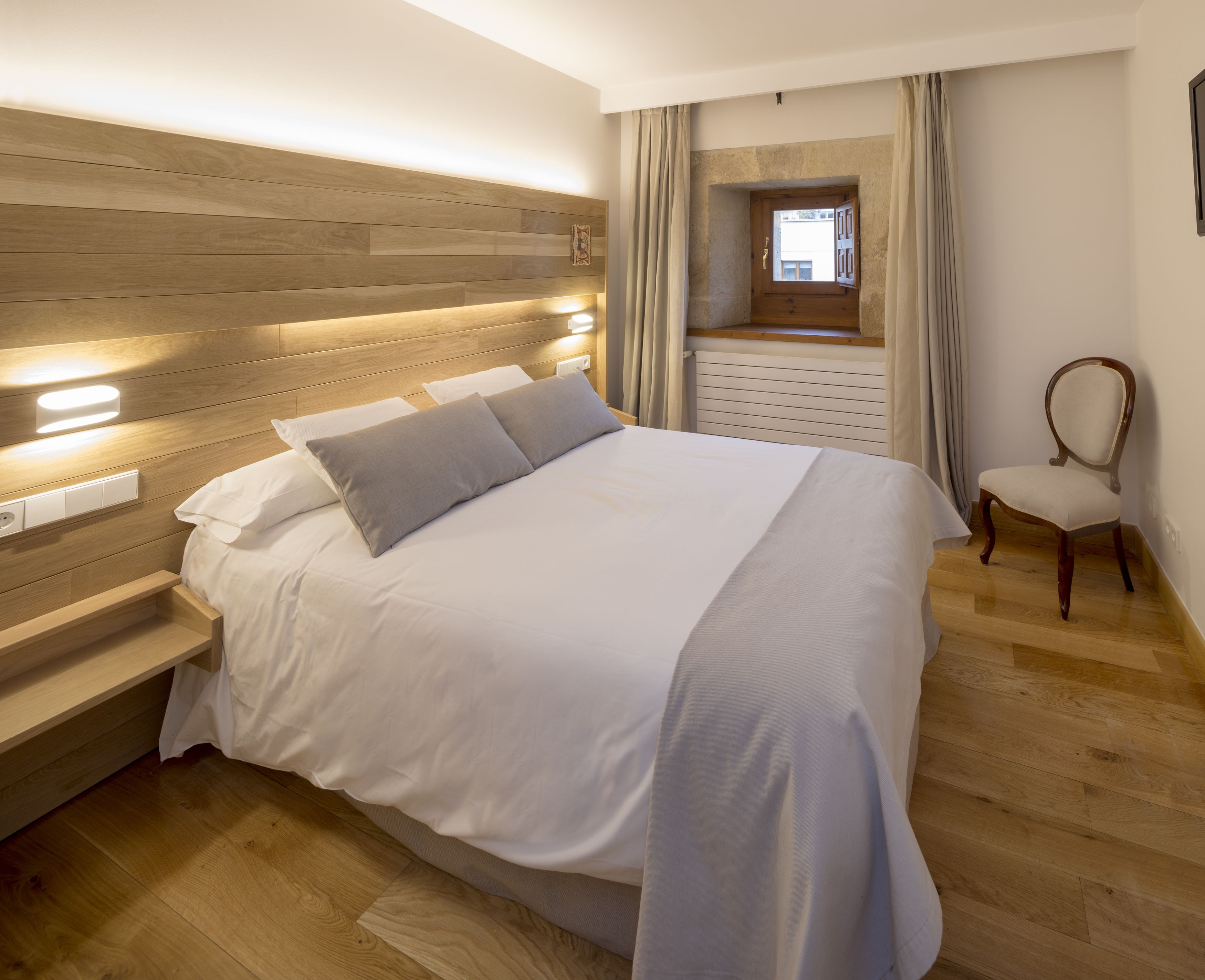 Una cama bien tendida con cabecera de madera, una mesita de noche con lámpara y una silla en una habitación de hotel con suelo de madera y ventana con cortinas.