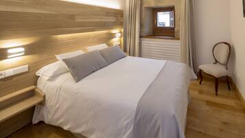 Una cama bien tendida con cabecera de madera, una mesita de noche con lámpara y una silla en una habitación de hotel con suelo de madera y ventana con cortinas.