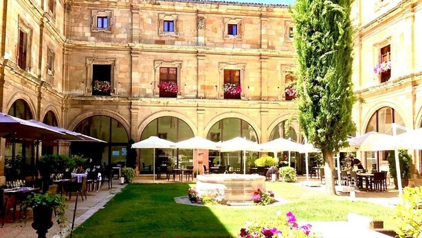 A courtyard with outdoor seating, a fountain, and a building with arched windows and balconies. - Hotel Real Colegiata San Isidoro (León)