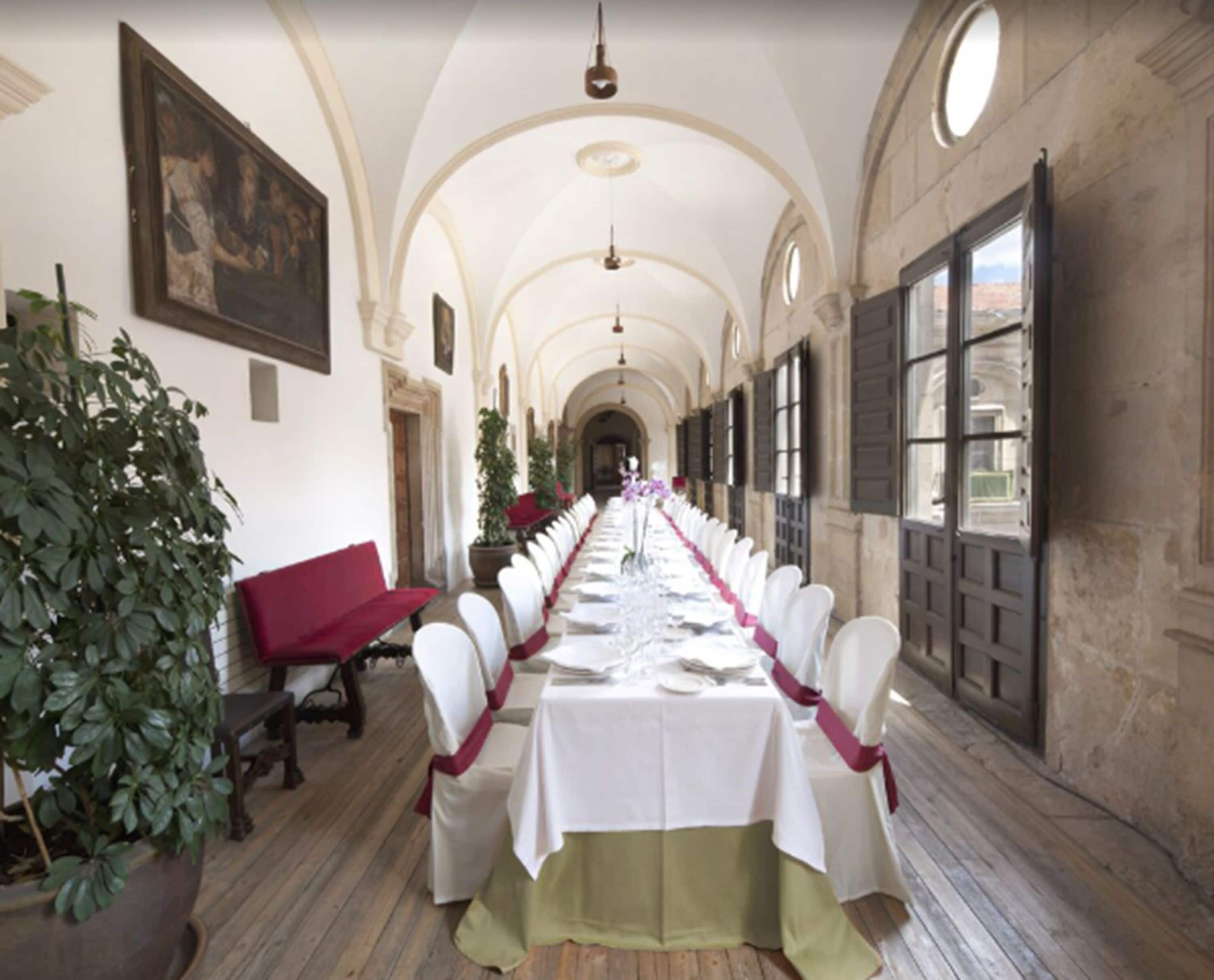 A long banquet table set for a formal event, with white chairs and a tablecloth, in a room with arched ceilings and large windows.