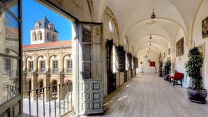 A corridor with arched ceilings, a view of a building with a tower, and potted plants along the walkway. - Hotel Real Colegiata San Isidoro (León)