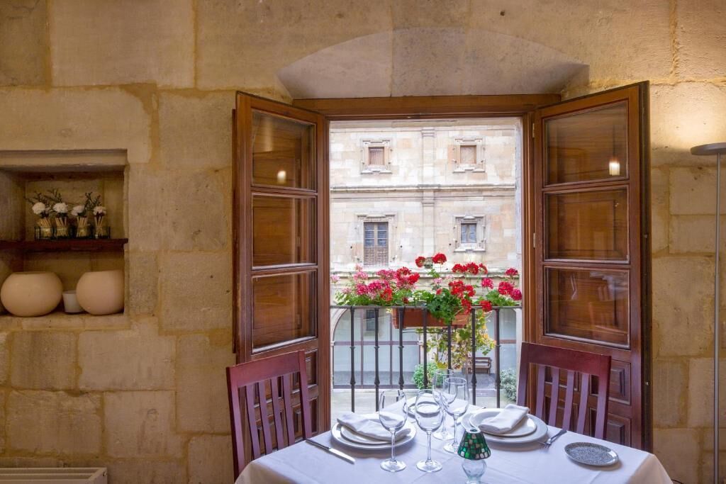 Un comedor con una mesa dispuesta para dos, un balcón con flores y vistas a un edificio.