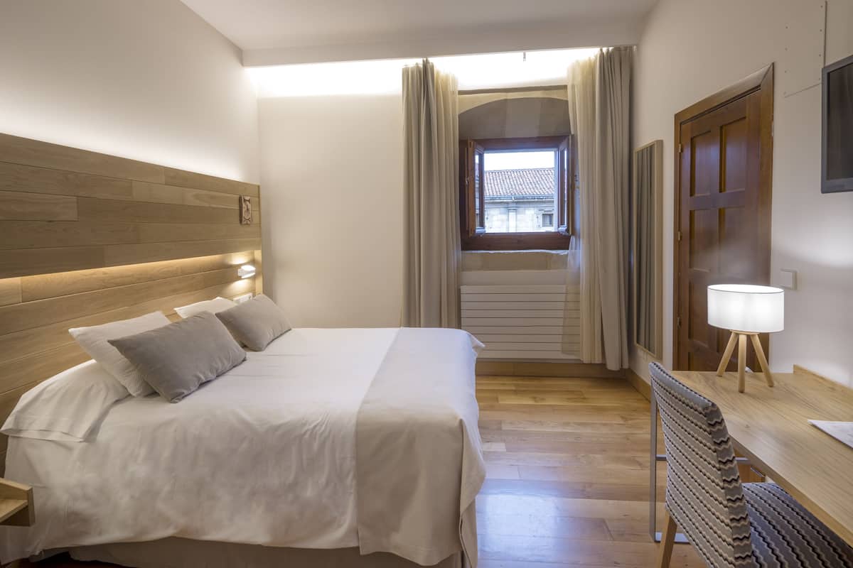A neatly arranged bedroom with a wooden headboard, a bed with white linens, a desk with a lamp, and a window with curtains.
