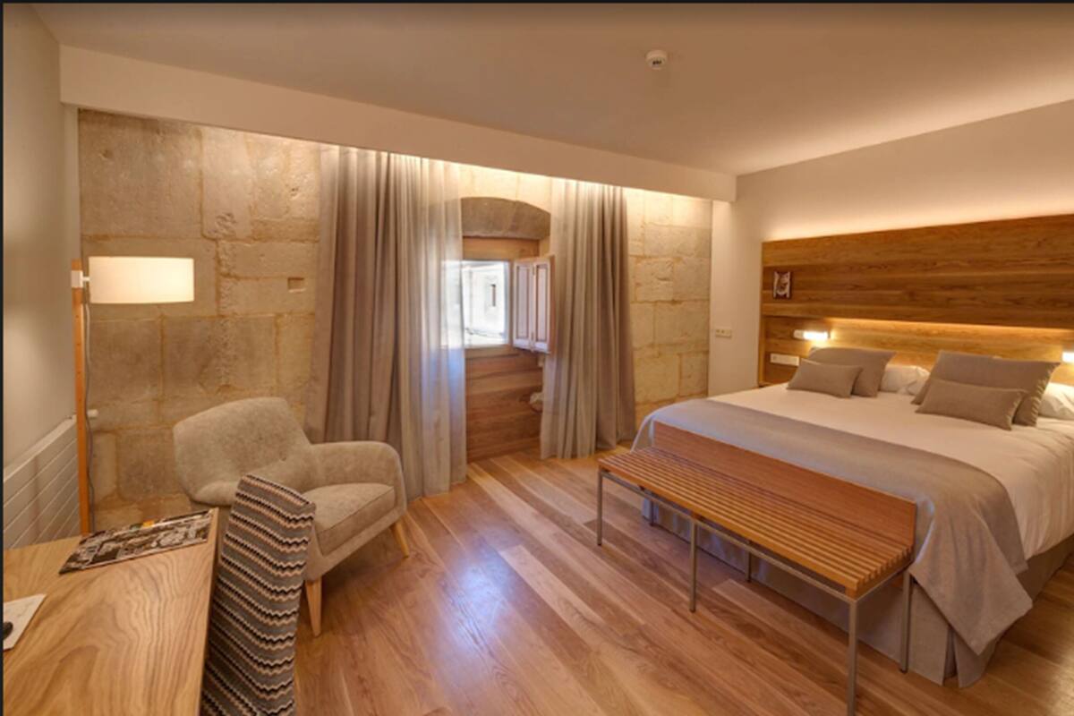 A modern bedroom with a wooden headboard, a large bed, a wooden bench, a chair, and a desk with a lamp.