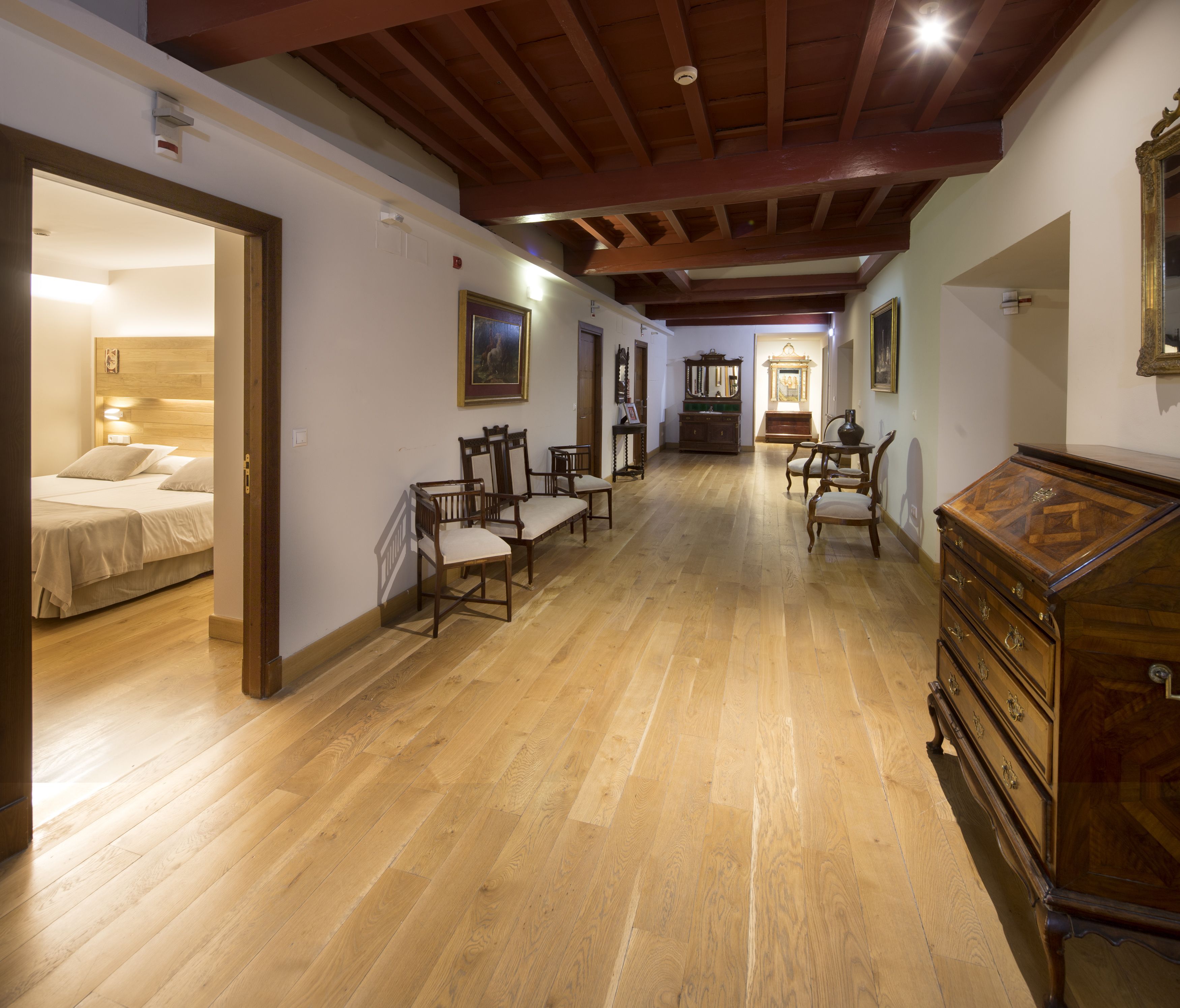 A spacious room with wooden flooring, a bed, several chairs, a dresser, and a hallway leading to another room.