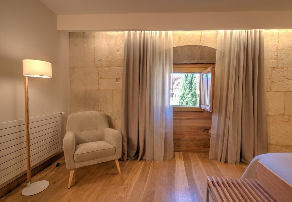 Una habitación con suelo de madera, un sillón beige, una lámpara de pie y una ventana con cortinas.