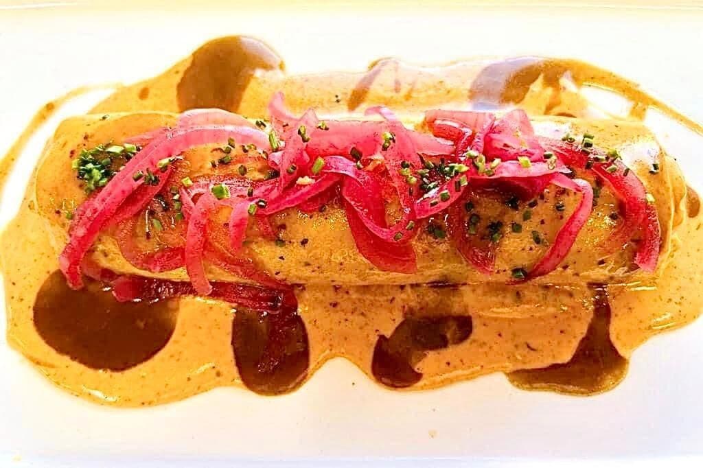 Un plat nappé d’une sauce crémeuse, garni d’oignons rouges finement tranchés et d’herbes fraîches, le tout présenté dans une assiette blanche.