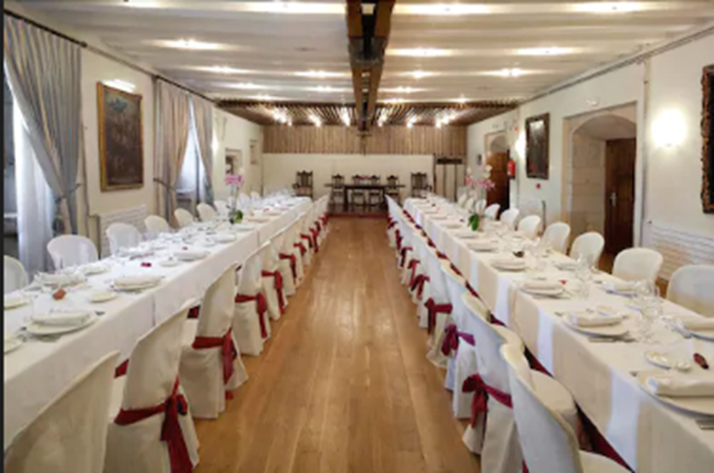 Una sala da banchetto con lunghe tavole imbandite per un evento formale, tovaglie bianche e sedie decorate con nastri rossi.