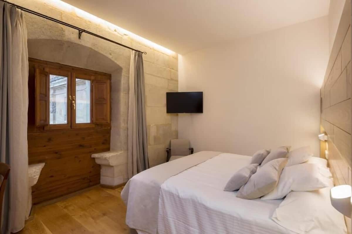 A bedroom with a large bed, a wooden bench, a television, and a stone wall.