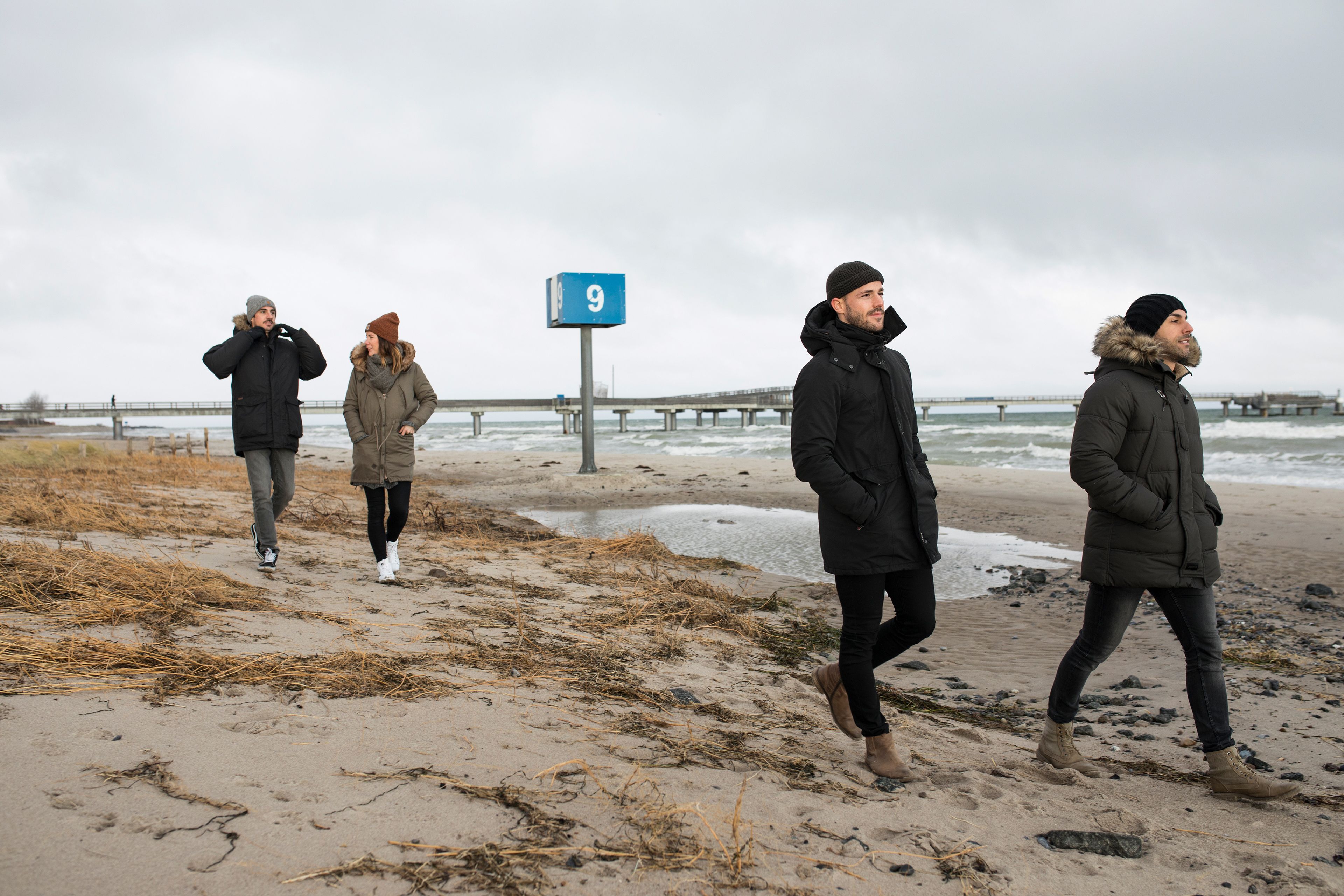 Una spiaggia con un molo sullo sfondo, un cartello con il numero 9 e persone che passeggiano lungo la riva.