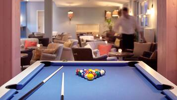 Une table de billard avec des billes au centre d’une pièce comprenant plusieurs coins salon et une personne à l’arrière-plan.