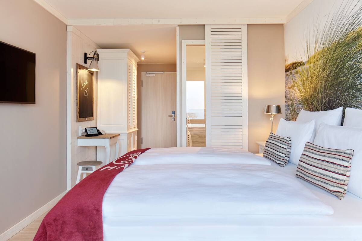 A bedroom with a large bed, a desk, a chair, a TV, and a door leading to another room.
